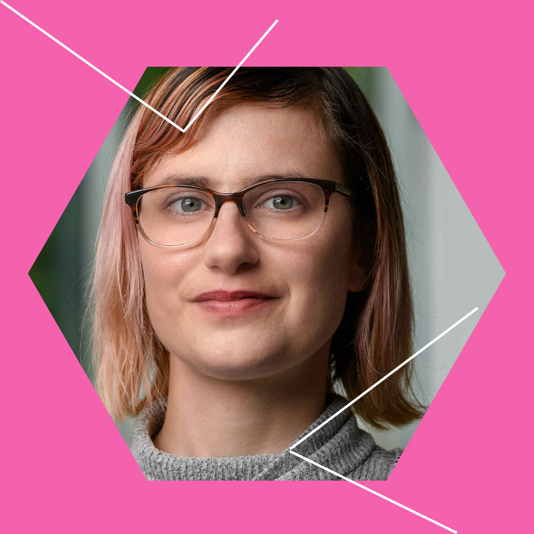 A hexagon frames Amy Reid's light skinned face with a slight smile, glasses and pink hair. The background is a hot pink with a few white angled lines.
