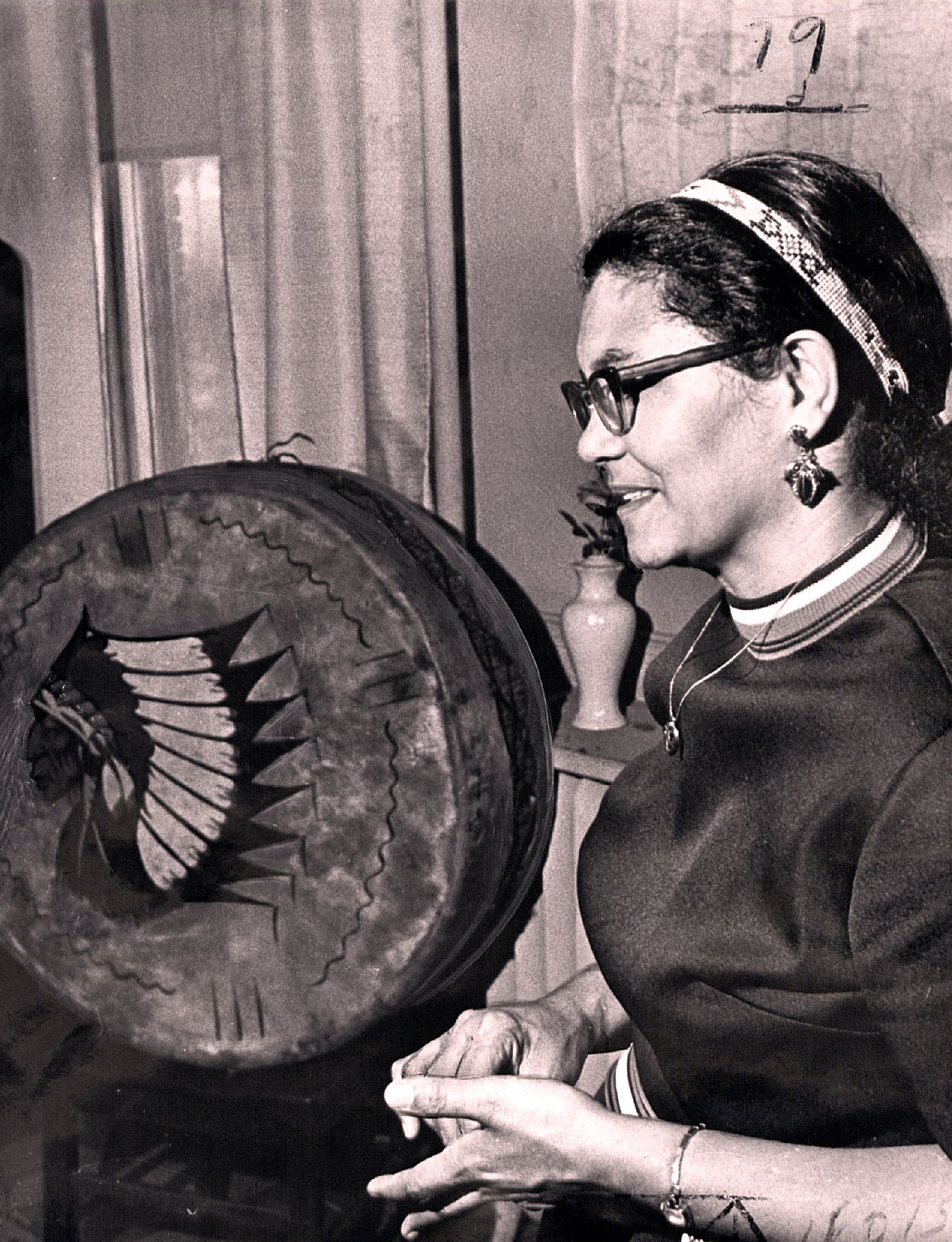 A black and white profile of Elizabeth Locklear with a large animal hide drum in the background. Elizabeth is in mid sentence clasping her hands with a beaded headband. Painted on the drum is the profile of a person wearing a warbonnet.