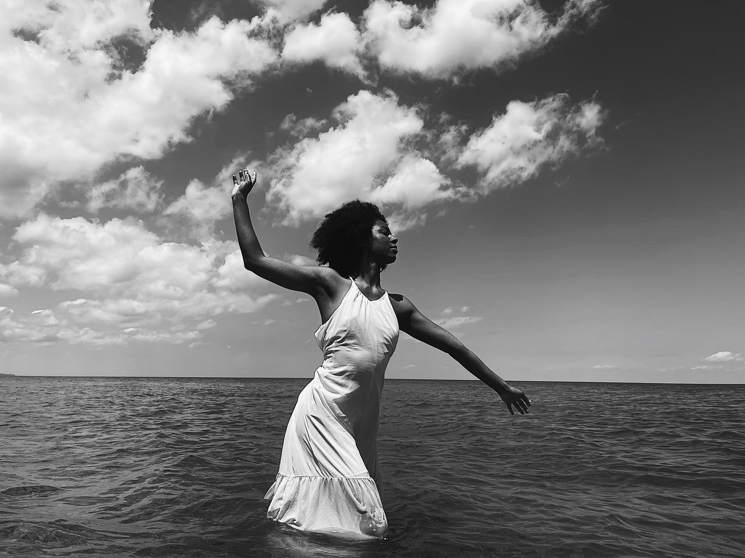 A black and white image of a medium dark skin tone person standing in the ocean. Their right arm is reaching back and up towards the cloud filled sky, their left arm reaches down and out towards the water. Their eyes are closed serenly.