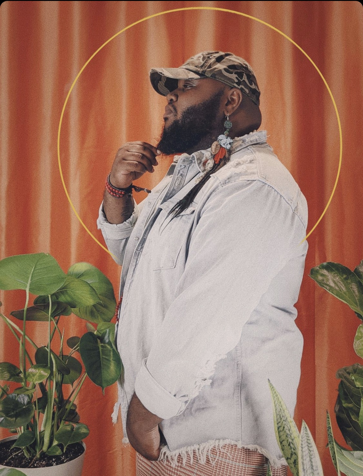 A profile portrait of a dark skinned person lightly holding the tip of their beard wearing a grey jean button down, long dangly earring and army print baseball cap. They are surrounded by house plants and a rich orange curtain hangs behind them