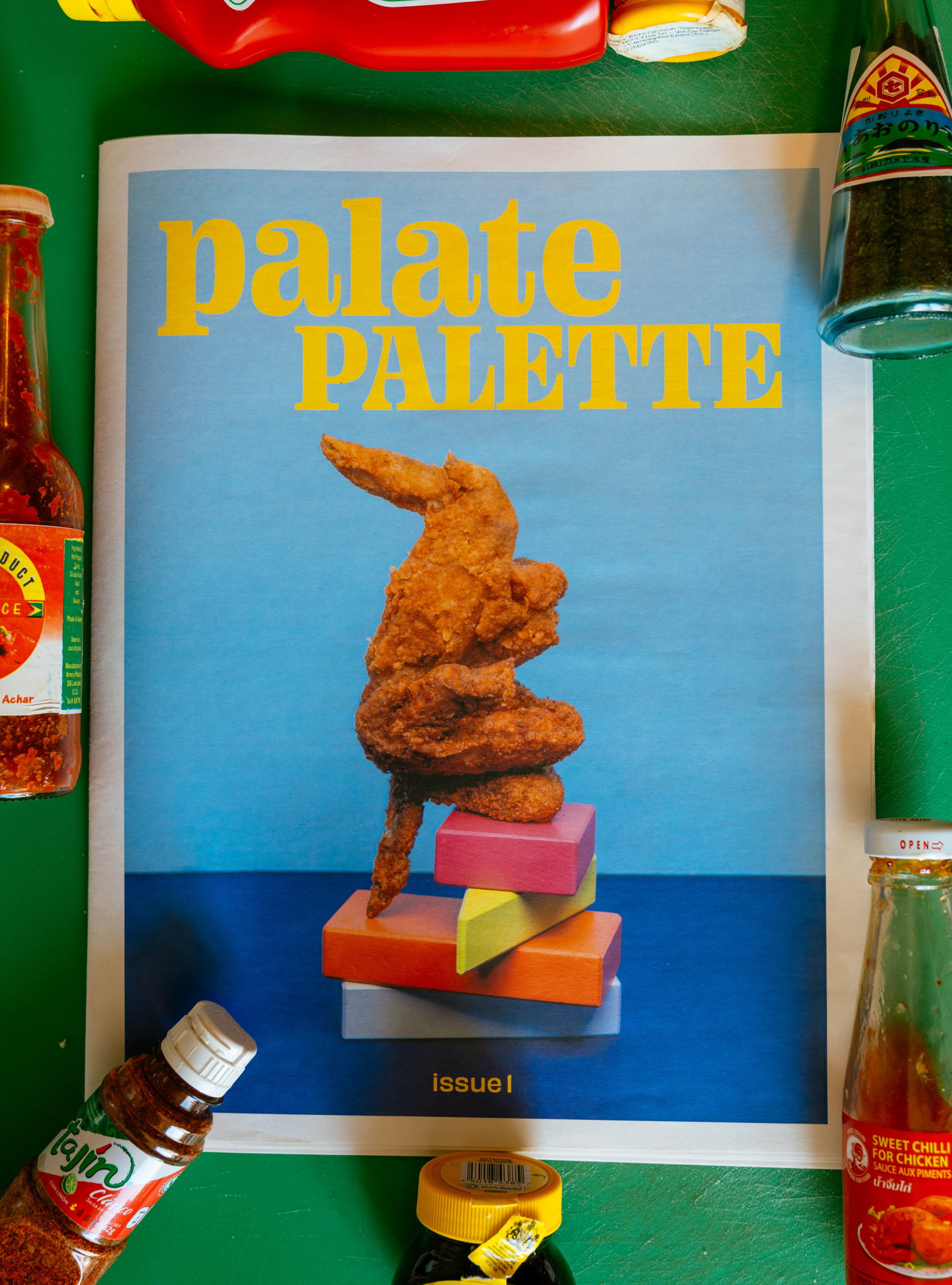 A piece of fried chicken balances on brightly colored stacked wooden blocks on the cover of the magazine Palate Palette. Hot sauce bottles surround the magazine.