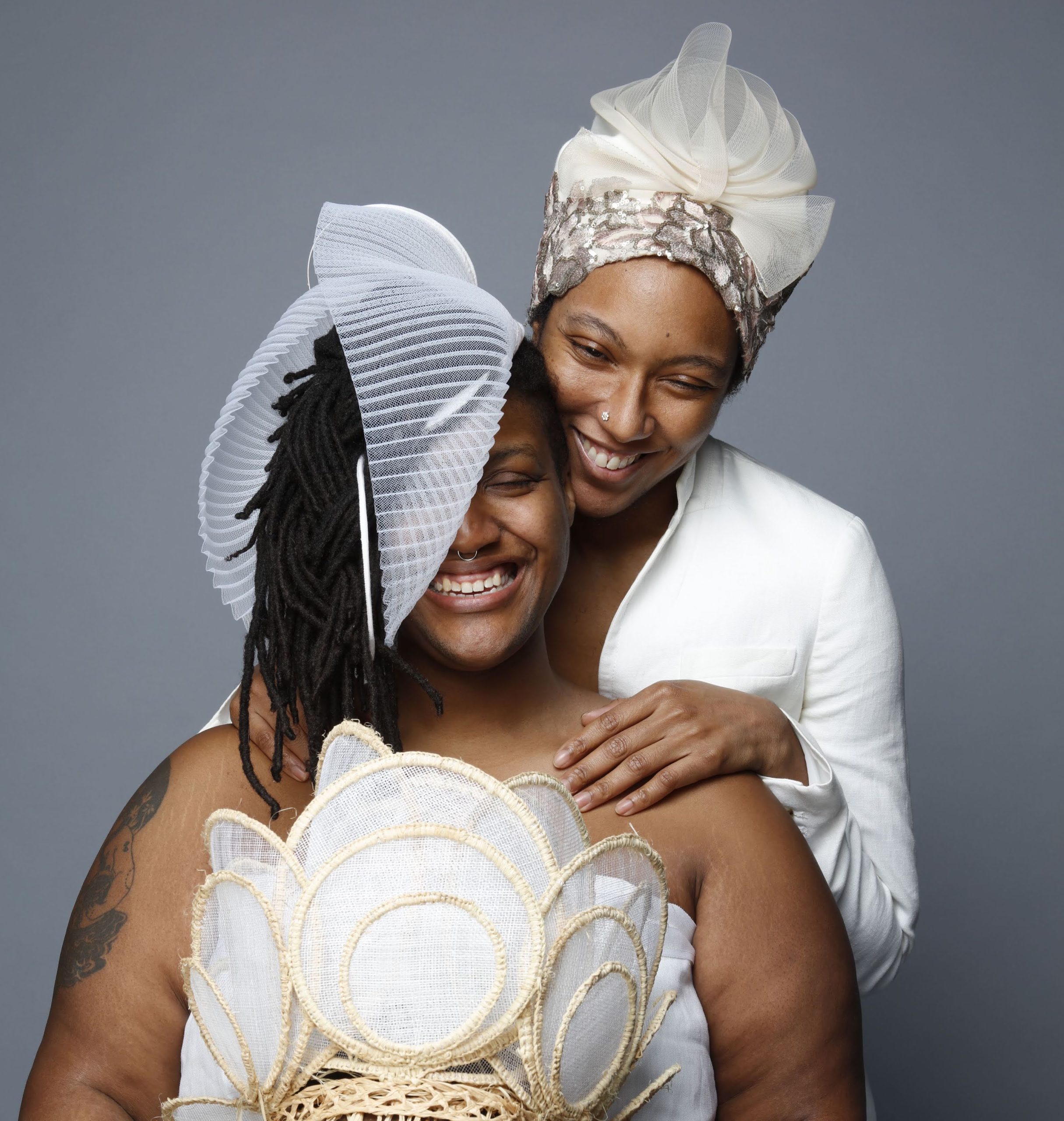 Two medium dark skinned people smile brightly, their eyes crinkling from laughter. Omolara stands behind Najee pressing O's cheek to Najee's head. They are adorned with white and cream sculptural head wear.