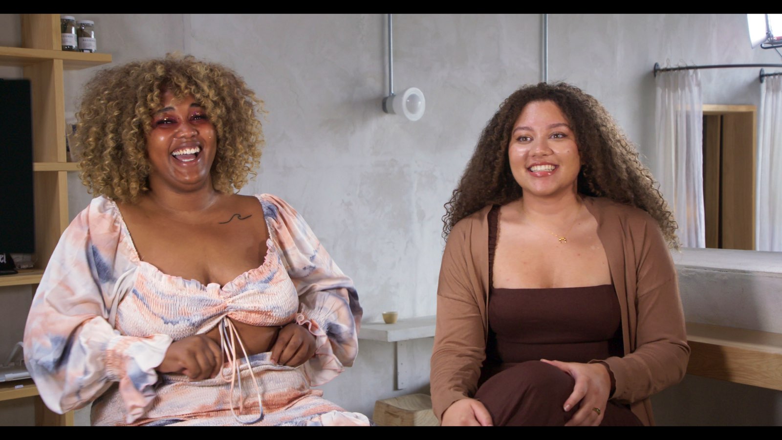 Two women with long curly hair sit on stools and laugh.